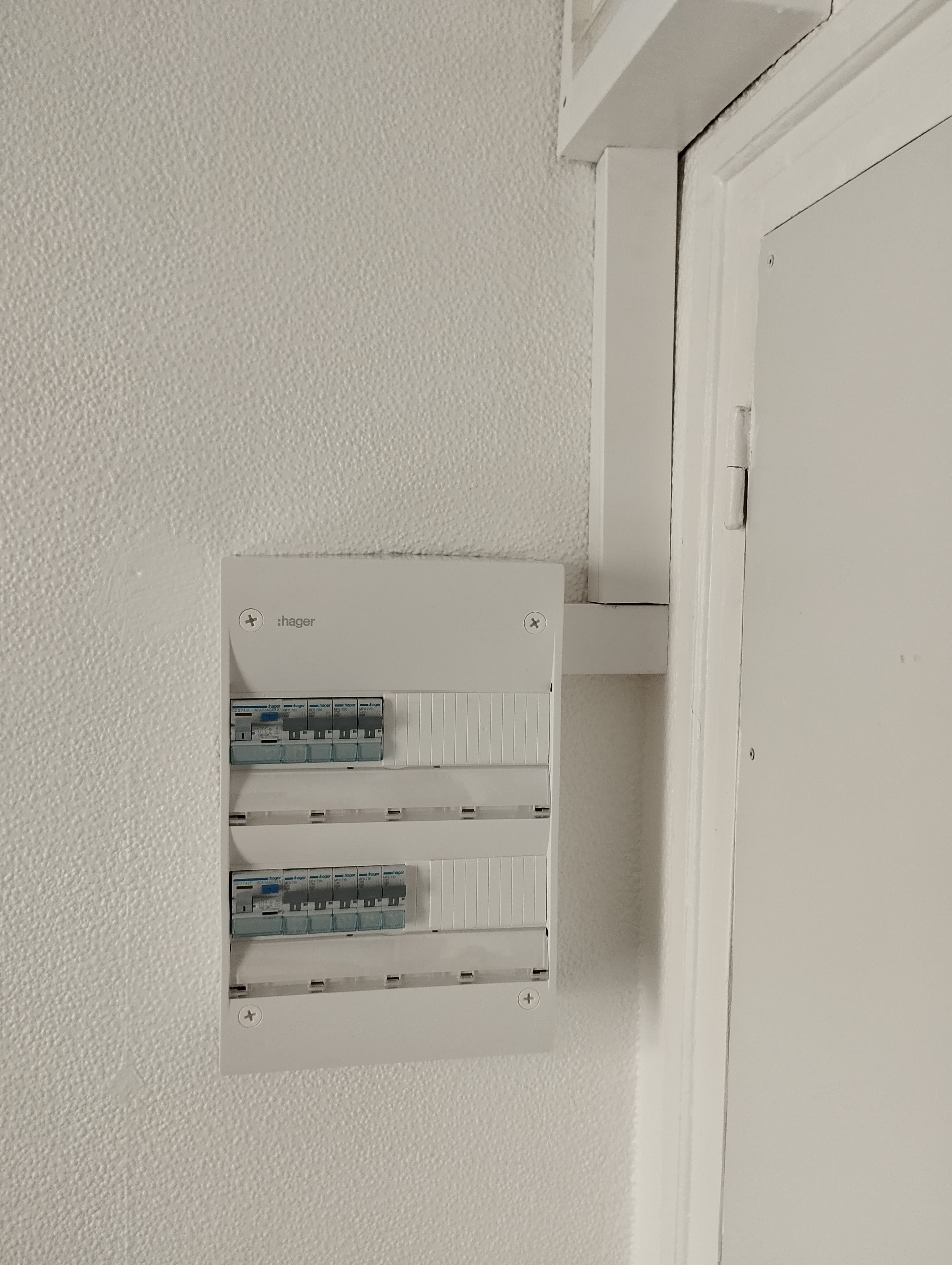 Tableau électrique blanc Hager avec plusieurs disjoncteurs fixé sur un mur blanc texturé.