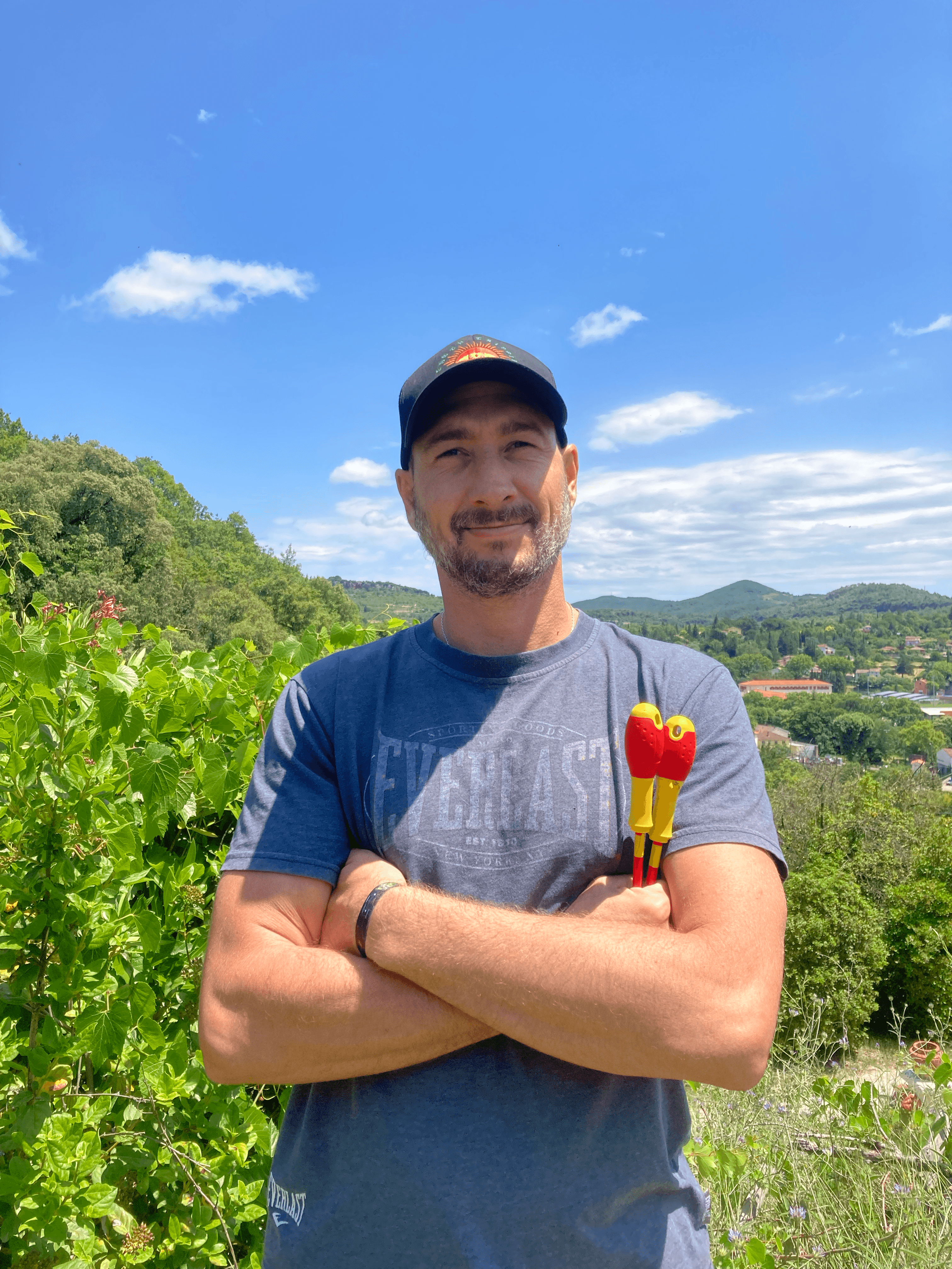 Homme barbu avec casquette, bras croisés tenant des tournevis devant un paysage de collines verdoyantes.